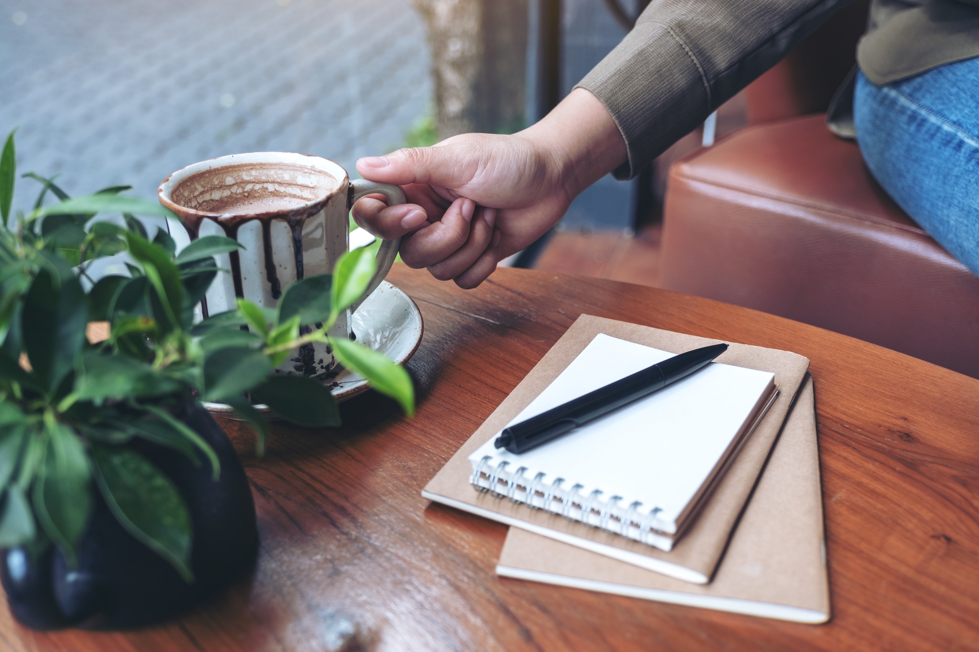 Closeup image of a hand grabbing a cup of hot coffee with notebooks on wooden table